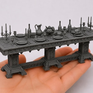 May include: A miniature grey resin dining table set featuring intricate gothic architectural details on the base. The tabletop is arranged with tiny plates, goblets, bottles, a pitcher, a fruit bowl, and candelabras, all rendered in a uniform matte grey finish.