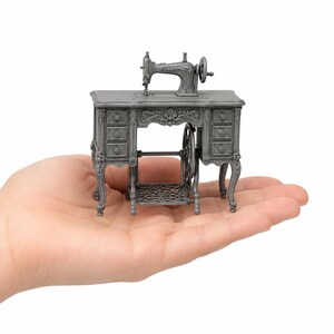 May include: A miniature silver-toned metal sewing machine mounted on a detailed desk with drawers, ornate carvings, and a foot pedal mechanism. The small-scale replica features a hand wheel on the side and a treadle base, showcasing intricate relief patterns.