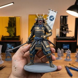 Peut inclure: Figurine détaillée d'un guerrier samouraï, tenant un katana et une bannière avec un emblème circulaire. La figurine est peinte en or, noir et bleu, et repose sur une base grise. L'arrière-plan comprend des équipements et des outils d'impression 3D.