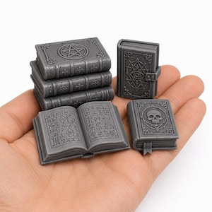 May include: A set of miniature gray resin books featuring intricate embossed details. The collection includes a stack of three books with a pentagram cover, an open book displaying runic symbols, a book with an eye emblem, and a skull-adorned book.