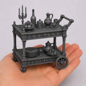 Puede incluir: Un carrito de servicio gris en miniatura con dos niveles y detalles ornamentados. El estante superior contiene un candelabro, una botella, un tarro con tapa, una jarra y un cuenco de fruta. El estante inferior contiene platos y una tetera.