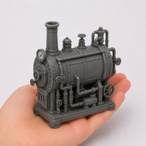 May include: A small, gray, three-dimensional printed model of a steam engine boiler. The object features a tall chimney, a circular gauge on the front, multiple pipes, valves, and wheels, all mounted on a rectangular base with rivet details.