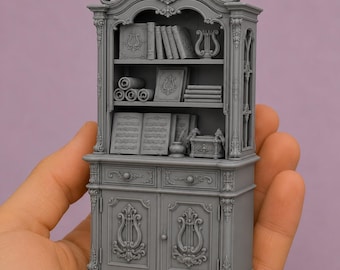 Könnte beinhalten: Ein grauer Miniaturschrank mit verzierten Schnitzereien, zwei Schubladen und zwei Türen mit Leier-Motiven. Die offenen Regale zeigen verschiedene Bücher, Schriftrollen, ein kleines Tintenfass mit Feder und eine dekorative Box in Grau.