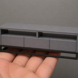 May include: A miniature gray entertainment center with three drawers and an open shelf. The cabinet has a black metal frame and is held in a person's hand. The piece is designed for a dollhouse or miniature setting.