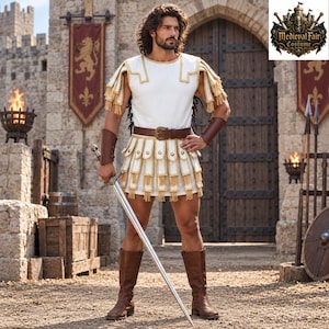 May include: A white tunic with gold fringe trim, a brown leather belt with a buckle, and matching brown leather bracers and knee-high boots. The costume includes a layered skirt with gold accents and a metal sword. The logo reads Medieval Fair Costume.
