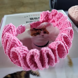 May include: A bright pink crocheted collar featuring a ruffled, wavy texture throughout the band. The accessory includes a small white plastic clasp closure. The design is constructed from yarn with a consistent, dense stitch pattern.