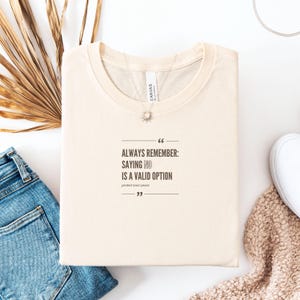 Könnte beinhalten: Cremefarbenes T-Shirt mit dem Text "ALWAYS REMEMBER: SAYING NO IS A VALID OPTION" in Schwarz. Das Shirt liegt gefaltet auf einer weißen Oberfläche, mit einer blauen Jeans und einem weißen Schuh.