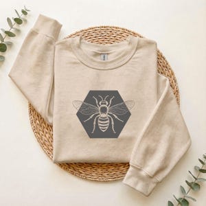 Könnte beinhalten: Ein beigefarbenes Sweatshirt mit Rundhalsausschnitt und einer dunkelgrauen Sechseck-Grafik auf der Brust. Im Inneren des Sechsecks befindet sich eine detaillierte weiße Strichzeichnung einer Biene mit ausgebreiteten Flügeln und Rippbündchen.