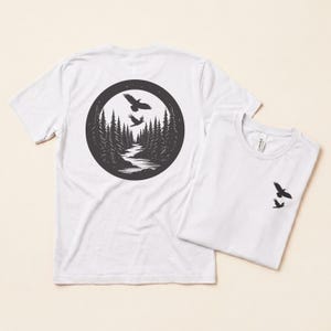 Könnte beinhalten: Ein weißes kurzärmeliges T-Shirt mit einer großen kreisförmigen schwarzen Grafik auf der Rückseite, die einen Wald, einen gewundenen Fluss und zwei fliegende Vögel zeigt. Eine kleinere Version der Vögel ist auf der linken Brustseite gedruckt.
