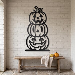 May include: A black metal wall art piece featuring a vertical stack of three carved pumpkins. The top pumpkin has a triangular nose and eyes, the middle has simple vertical slits, and the bottom has a wide, curved, toothy grin.