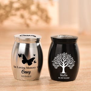 May include: Two small metal cremation urns, one silver and one black. The silver urn features a butterfly graphic and the text In Loving Memory Of Casey 8.13.53-9.20.21. The black urn features a tree graphic and the text Muffin 04.1967-06.2026.