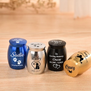 May include: Four small metal cremation urns in blue, silver, black, and gold. Each urn is engraved with a name, including Sadie, Ruby, Oscar, and Casey, along with dates, paw prints, hearts, or butterfly designs and the text In Loving Memory.