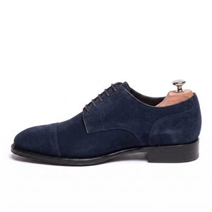 Könnte beinhalten: Ein marineblauer Oxford-Schuh aus Wildleder mit Zehenkappe, dunkelbraunen Schnürsenkeln und einem schwarzen Absatz. Der Schuh ist mit einem hölzernen Schuhspanner versehen, um seine Form zu bewahren, und zeigt ein klares Profil mit glatter, matter Textur.