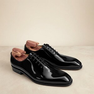 May include: A pair of black patent leather oxford shoes with a high-gloss finish, featuring thin black laces, a rounded toe, and a low heel. Each shoe contains a wooden shoe tree insert that is visible at the opening of the footwear.