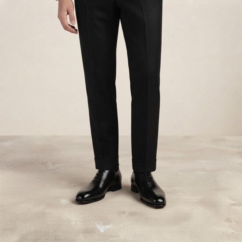 May include: A pair of black leather oxford shoes with a polished finish, featuring thin black laces and a rounded toe, worn with tailored black trousers that have a clean, straight-leg cut and a visible hem at the bottom of the pant legs.