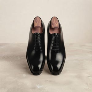 May include: A pair of black leather oxford shoes with a polished finish, featuring thin black laces and wooden shoe trees inserted into the openings. The shoes have a rounded toe design and a clean, smooth exterior surface with visible stitching.