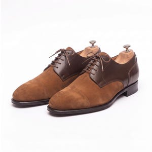 Könnte beinhalten: Ein Paar Oxford-Schuhe mit Zehenkappe im zweifarbigen Design mit hellbraunem Wildleder vorne und dunkelbraunem Glattleder hinten. Die Schuhe verfügen über dünne braune Schnürsenkel und in jedem Schuh stecken hölzerne Schuhspanner.
