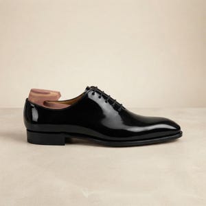 May include: A black patent leather oxford shoe with a closed lacing system and thin black laces. The shoe features a low heel and a smooth, polished finish. A wooden shoe tree is inserted into the heel area, showing a natural light brown grain.