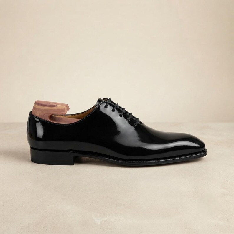 May include: A black patent leather oxford shoe with a closed lacing system and thin black laces. The shoe features a low heel and a smooth, polished finish. A wooden shoe tree is inserted into the heel area, showing a natural light brown grain.