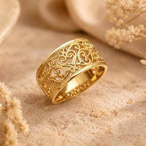 May include: A wide gold ring featuring an intricate openwork filigree pattern of swirling scrolls and heart-like motifs. The band has a polished finish with a consistent width throughout, showcasing detailed metalwork that creates a lace-like appearance.