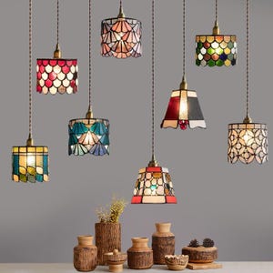 May include: A collection of eight hanging pendant lamps with stained glass shades in various shapes and colors, including red, blue, green, and white patterns. Each lamp features a brass-toned fixture and hangs from a twisted brown fabric cord.