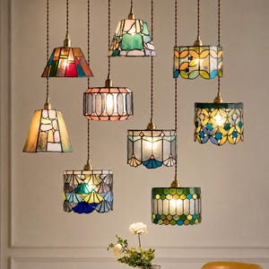 May include: A collection of nine hanging pendant lamps featuring stained glass shades in various geometric shapes and colorful patterns. Each lamp is suspended by a brown twisted cord and emits a warm light through the multi-colored glass panels.