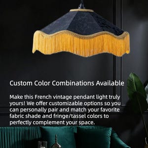 Op de afbeelding: Een hanglamp met een zwarte stoffen kap in een zeshoekige vorm, geaccentueerd met een goudkleurige gevlochten rand aan de bovenkant en een lange, golvende goudkleurige franje langs de onderkant. De lamp hangt aan een zwart snoer en armatuur.