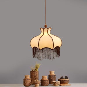May include: A pendant light fixture featuring a bell-shaped, off-white fabric shade with dark brown trim and a long, dark brown fringe along the bottom edge. The lamp is suspended by a thin cord from a small, round, dark wood ceiling mount.