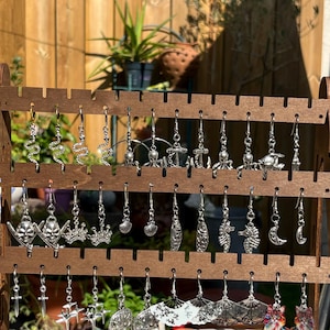 Peut inclure: Un présentoir à boucles d'oreilles en bois présentant diverses boucles d'oreilles argentées. Les motifs comprennent des serpents, des ancres, des crânes, des couronnes, des coquillages, des hippocampes, des lunes, des croix, des éventails et des boucles d'oreilles en forme de chat coloré.
