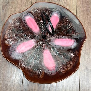 May include: A decorative serving dish with a brown and pink floral design. The dish has a black handle and a unique, textured pattern. The rim is a darker brown, and the pink sections are in a petal shape.