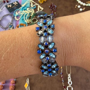 May include: A stretch bracelet featuring three floral-shaped metal links in a dark gunmetal tone, each adorned with light blue and dark blue faceted crystals, separated by clusters of translucent light blue round beads in a repeating pattern.