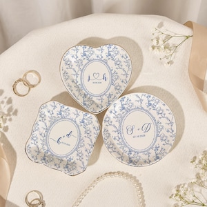 May include: Three porcelain dishes in heart, square, and round shapes. Each dish has a blue floral pattern and a central monogram with a date. Gold rims and a white background complete the design.