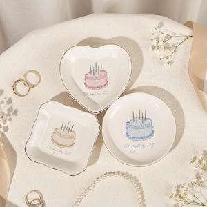 May include: Three white ceramic trinket dishes with silver rims. One heart-shaped dish features a pink cake with candles and the text Chapter 25. One square dish shows a beige cake with candles and text. One round dish has a blue cake and text.