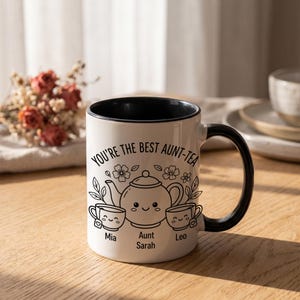 May include: A white ceramic mug with a black interior and handle, featuring a black line art illustration of a teapot and two teacups with smiling faces. The text reads: YOU'RE THE BEST AUNT-TEA, with the names Mia, Aunt Sarah, and Leo below.