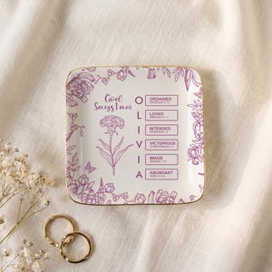 Peut inclure: Un vide-poche carré en céramique blanche avec un bord doré, orné d'illustrations florales violettes et du texte God Says I Am, du prénom OLIVIA, et de six mots encadrés : ORDAINED, LOVED, INTENDED, VICTORIOUS, IMAGE, et ABUNDANT avec versets.