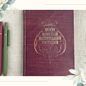 May include: A maroon hardcover book titled Levi Wilbur Pollard's Poems with gold embossed lettering and a lyre design on the front cover, accompanied by two matte finish pens in dark red and olive green colors placed vertically to the left.
