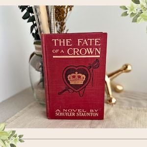May include: A red hardcover book titled The Fate of a Crown by Schuyler Staunton. The front cover features a gold-colored crown inside a heart shape, with a sword graphic behind it. The text is printed in a clear, gold-colored serif font.