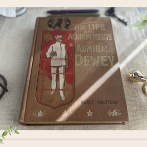 May include: A hardcover book titled The Life and Achievements of Admiral Dewey by Murat Halstead. The tan cover features a gold-toned illustration of a naval officer in uniform inside a red shield, with gold lettering and four white stars at the base.