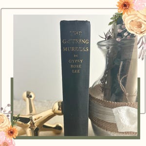 May include: A vintage hardcover book titled "The G-String Murders" by Gypsy Rose Lee. The book has a dark blue cover with gold lettering. A gold decorative object and a glass vase with floral accents are also in the image.