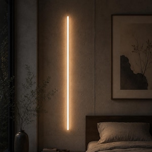 Puede incluir: Una lámpara de pared lineal y vertical que emite un brillo cálido y suave. La lámpara cilíndrica y delgada está colocada contra una pared de hormigón texturizado, proporcionando una iluminación ambiental a lo largo de la barra de luz.
