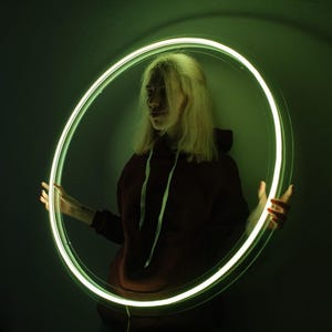 May include: A person is framed by a glowing green neon circle. The person wears a maroon hoodie with white drawstrings. The background is dark, with the neon circle providing the primary light source.