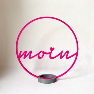 Könnte beinhalten: Ein leuchtend pinkfarbenes, kreisförmiges Dekoschild mit dem Wort moin in Schreibschrift und einem kleinen Herz über dem Buchstaben i. Der kreisförmige Rahmen wird von einem massiven grauen zylindrischen Sockel aufrecht gehalten.