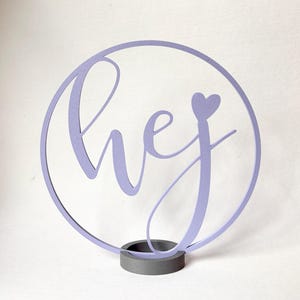 Könnte beinhalten: Ein hellviolettes, kreisförmiges Schild mit dem kursiven Schriftzug "hej" und einem kleinen Herzsymbol über dem Buchstaben "j". Das Schild ist auf einem massiven, dunkelgrauen zylindrischen Sockel befestigt und bildet ein freistehendes Dekorationsobjekt.