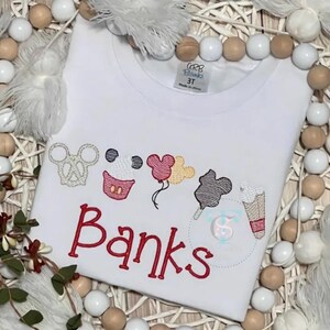 May include: White t-shirt featuring embroidered Mickey Mouse-themed designs, such as a pretzel, cupcake, balloons, candy floss, and ice cream. The name "Banks" is embroidered in red. The shirt is labelled "3T".