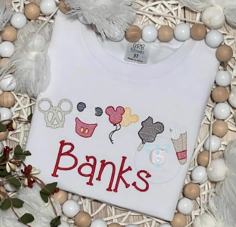 May include: White t-shirt featuring embroidered Mickey Mouse-themed designs, such as a pretzel, cupcake, balloons, candy floss, and ice cream. The name "Banks" is embroidered in red. The shirt is labelled "3T".