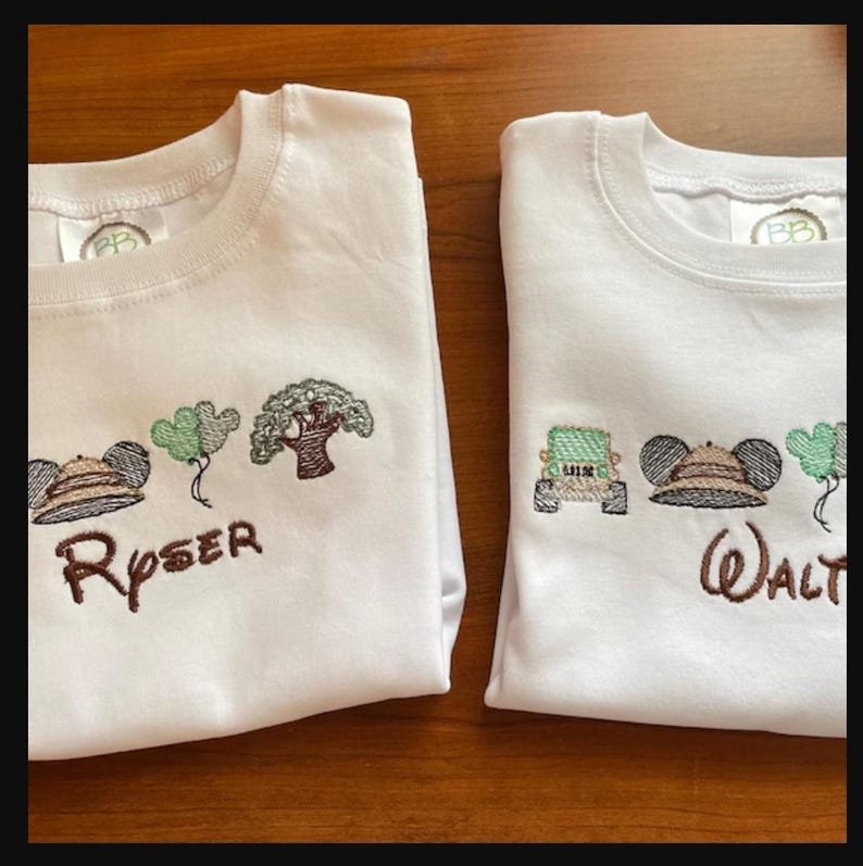 May include: Two white crew neck shirts with embroidered designs. One shirt features a safari hat, trees, and the name "Ryser". The other shirt has a jeep, safari hat, trees, and the name "Walt".