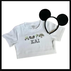 May include: White t-shirt with embroidered design and the name "KAI". The design includes a car, Mickey Mouse ears, and other elements. A black headband with large black mouse ears is also present.