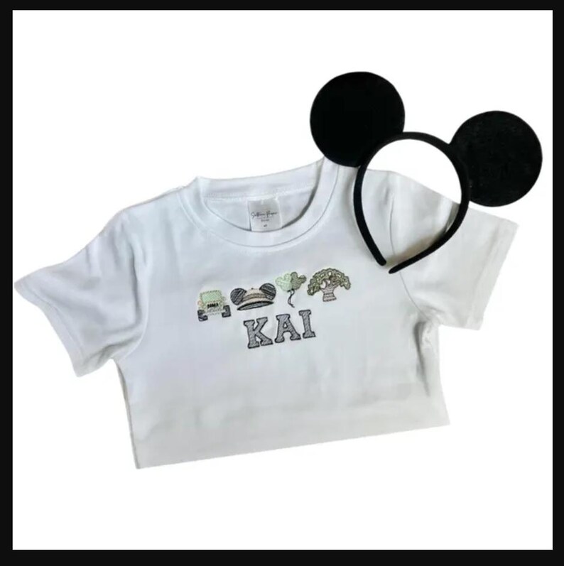 May include: White t-shirt with embroidered design and the name "KAI". The design includes a car, Mickey Mouse ears, and other elements. A black headband with large black mouse ears is also present.
