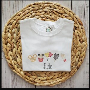 May include: White t-shirt with embroidered cartoon character designs and the name "Jude" in black script. The shirt has a crew neck and short sleeves. The shirt is displayed on a woven, circular placemat.