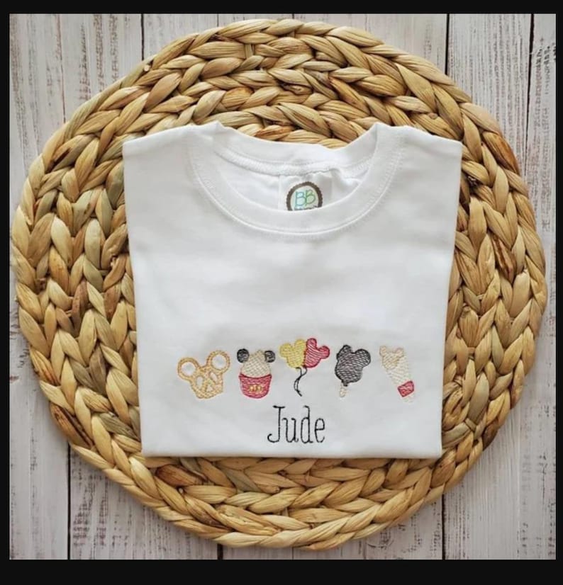 May include: White t-shirt with embroidered cartoon character designs and the name "Jude" in black script. The shirt has a crew neck and short sleeves. The shirt is displayed on a woven, circular placemat.